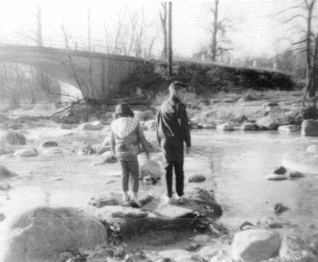 Blurry black and white photo of two children standing on boulders in a river.