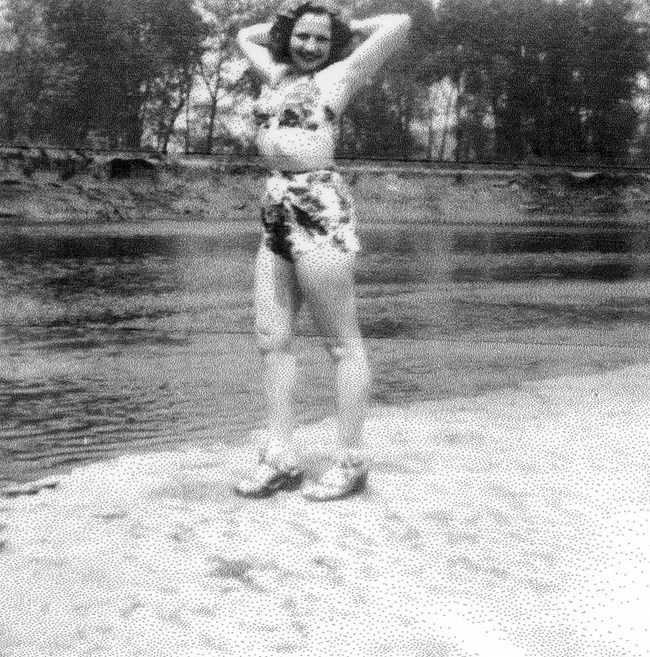 A smiling young woman with wavy hair stands along a riverbank with her arms behind her head. She wears a patterned bikini and heels.