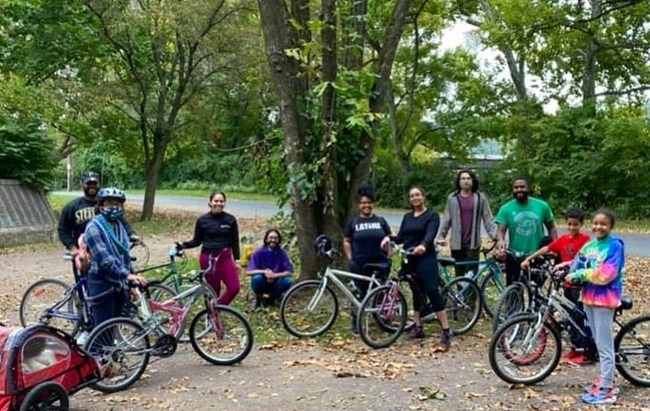 D&L Family Ten people of varied ages (adults and children) with bikes stand together in park environment with large tree behind