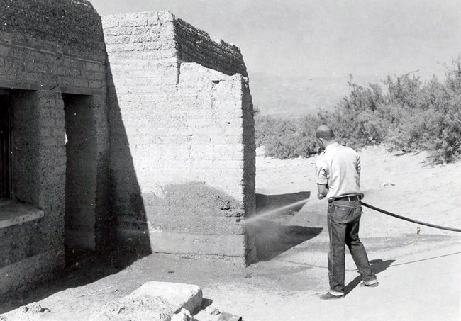 An NPS ranger washes down an Timbisha Shoshone adobe house. Even though the NPS was in charge of maintaining the buildings, they used their “neglect” to justify washing them away. April 1958. Man uses hose and blasts wall of adobe building with water.