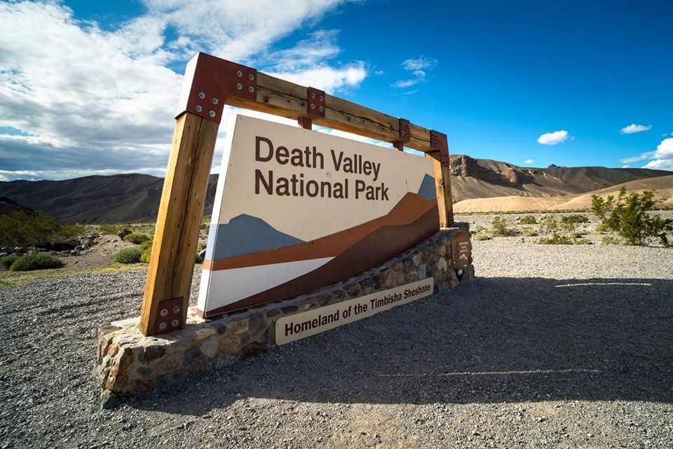 DEVA_Sign A large sign that reads "Death Valley National Park: Homeland of the Timbisha Shoshone" with a wooden border and stone foundation, with brown hills and a partly cloudy blue sky in the background.