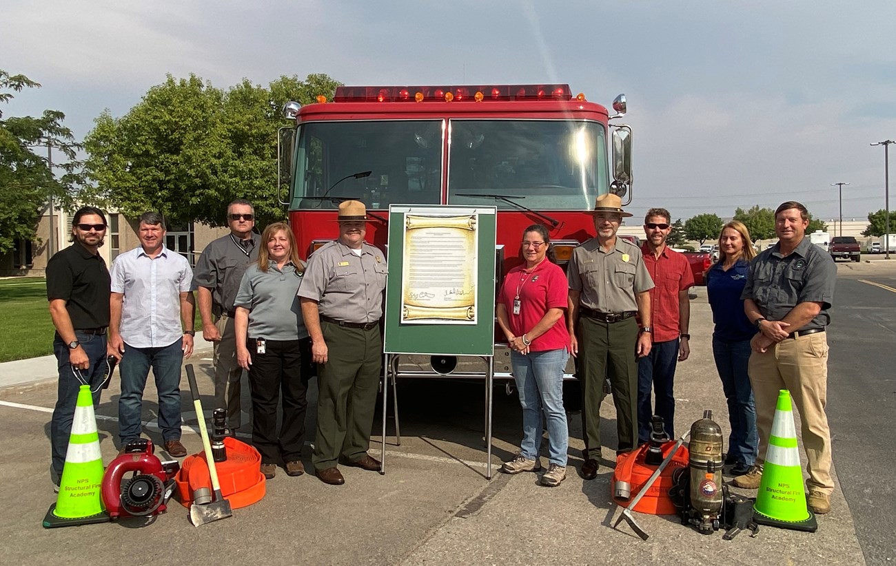 Division of Fire and Aviation gather for Fire Prevention Week Proclamation signing A group of people stand in front of a fire engine with a board holding a sign.