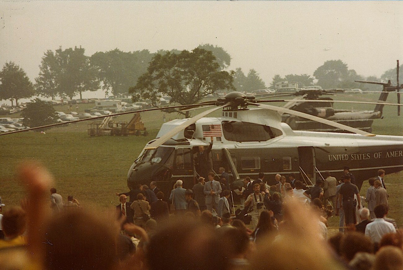 A helicopter lands on the grass near a crowd of people.