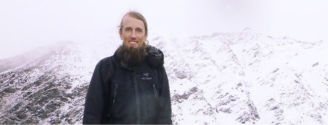 A bearded man dressed in black stands in front of a misty, snow-covered mountain.