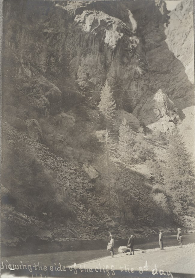 Pelton Expedition 1900 Men, probably J.E. Pelton, J.A. Curtis, M.F. Hovey, and E.B. Anderson, stand by the Gunnison River, in the Black Canyon of the Gunnison. One person points up at steep, tall canyon walls.