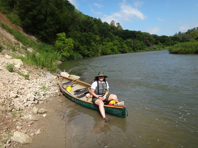 A canoer sitting on a canoe with a canyon wall to the left and river to the right. The sky is blue.