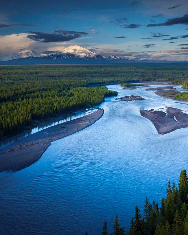 A deep blue river flows through a green, forested and mountainous landscape.