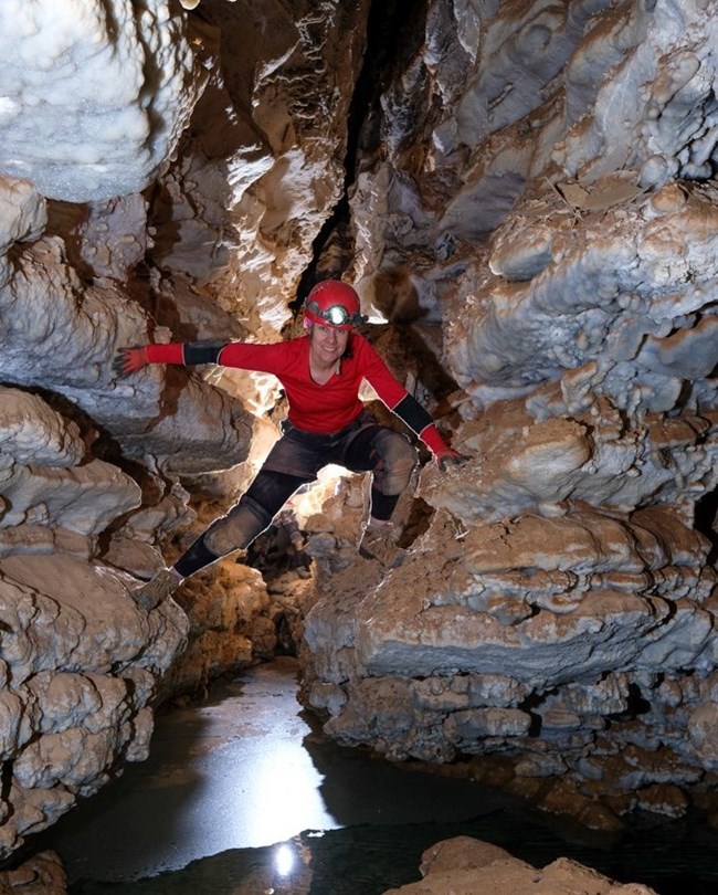 a caver standing on a ledge in a narrow cave passage over a shallow underground lake