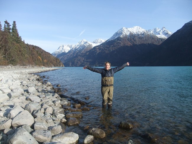 Lisa standing in a huge lake surrounded by tall, jagged peaks, holding her arms out in an expression of joy.