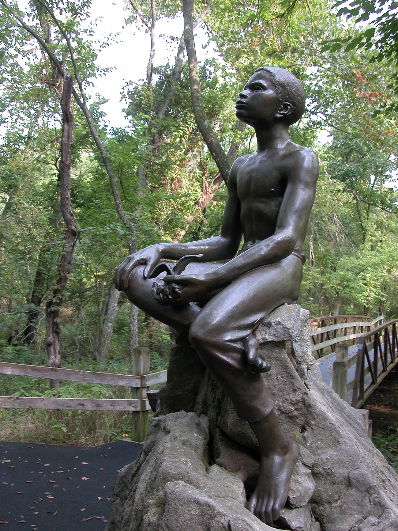 Bronze statue of George Washington Carver as a young child. He is holding a plant and sitting on a large boulder.