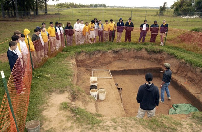 Jeff Girard stands in an excavation pit as school children gather behind fencing to listen.