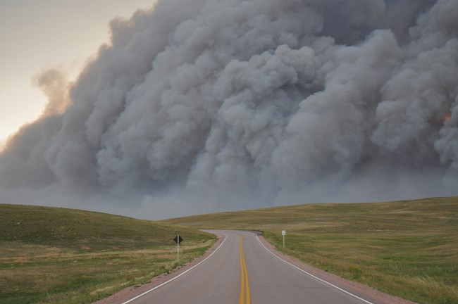an empty prairie highway with a dark ominous cloud of smoke rising in the distance covering most of the sky