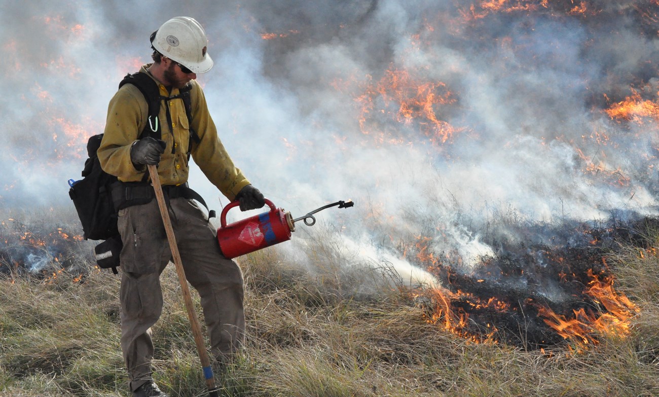 a wildland firefighter holding a drip torch in front of a smoldering grass fire in the prairie