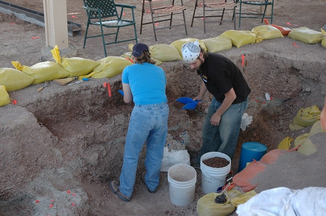 two paleontologists work in a large pit surrounded by tools and flags identifying fossils