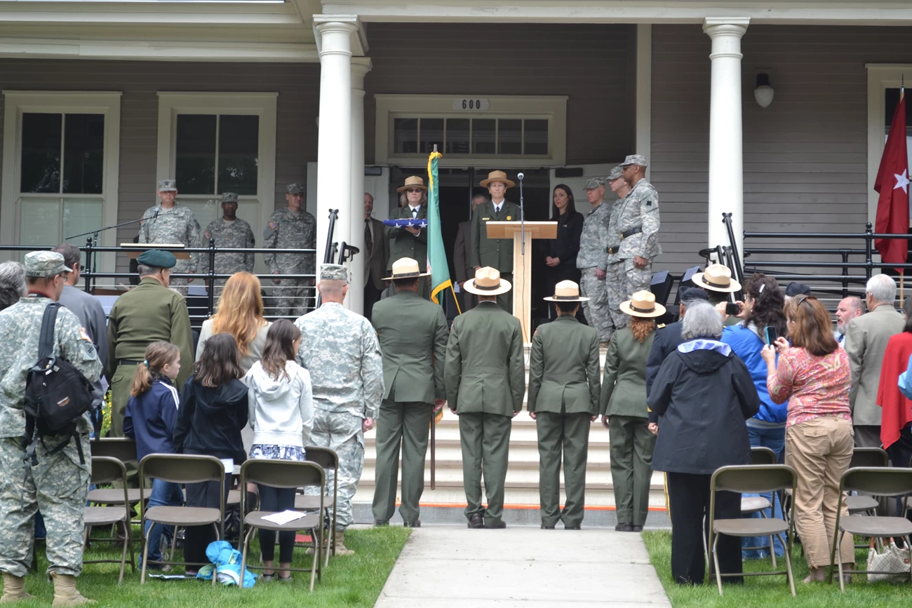 Post to Park 2 A row of National Park Service and US Army representative stand on the porch of a historic barracks building. One ranger is holding a folded flag. A line of National Park Service rangers stands at attention facing them.