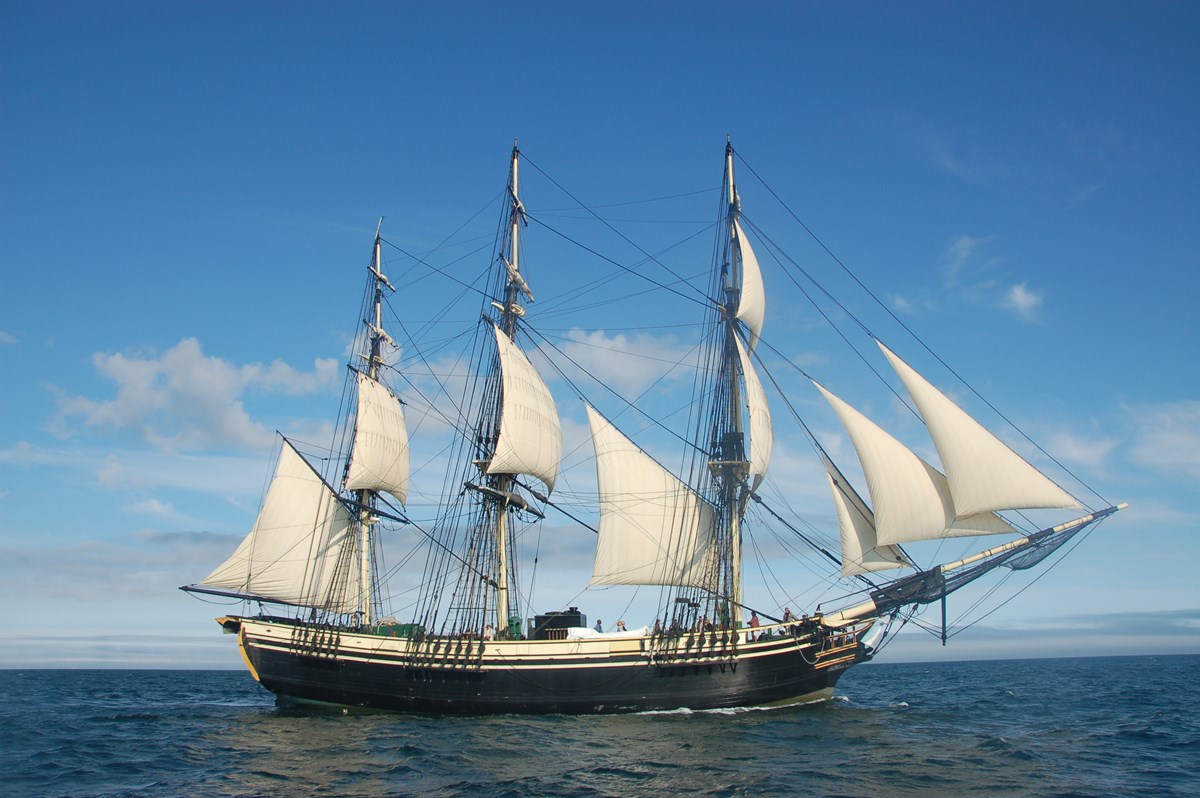 A large three-masted vessel with black hull and white sails travels through the water with no land in sight.