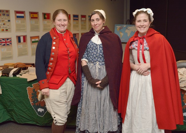 three women wearing living history clothing stand in front of a table indoors