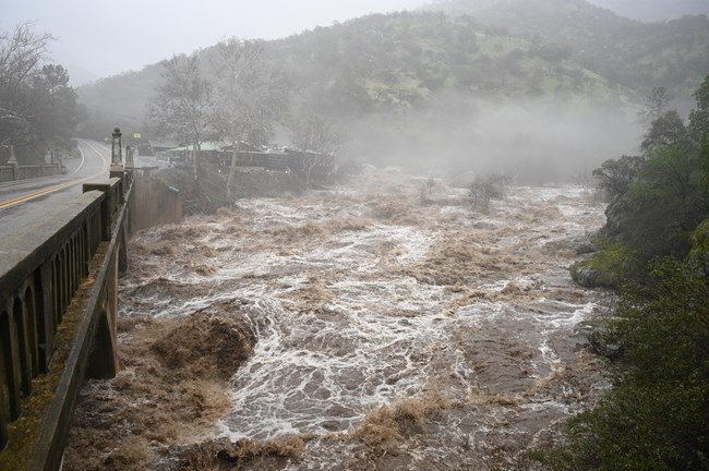 View from bridge of sediment-filled, high water flowing in river.