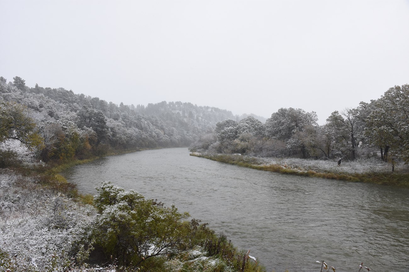 A river flows through a landscape of bluffs and snow is beginning to cover the trees
