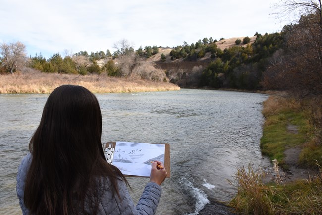 A woman stands drawing the river and cliffs in the distance.