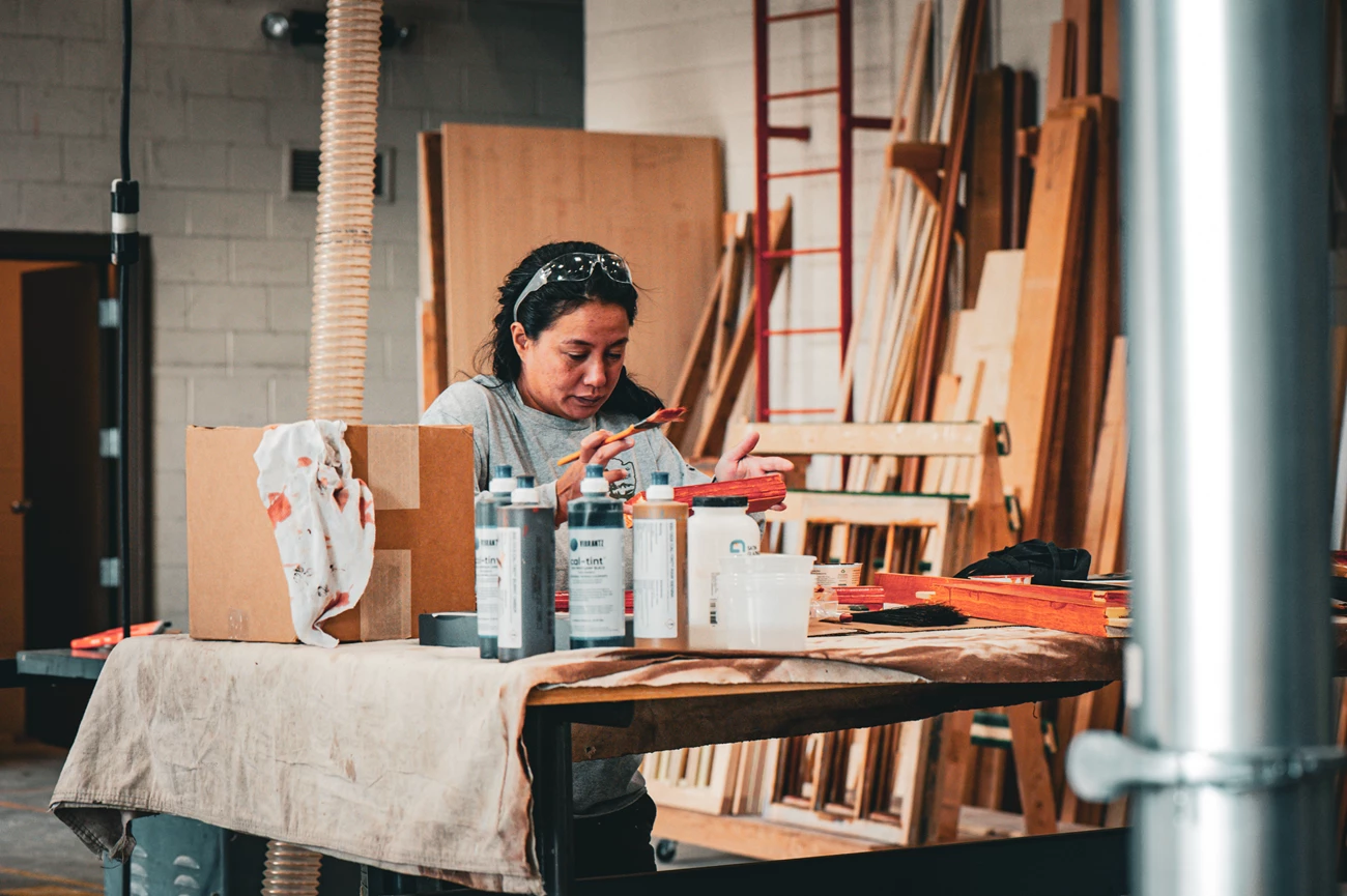 FOVA HPTC A woman in a National Park Service uniform applies stain to historic wood parts from a building. She has safety goggles up on her head. In front of her on a work table are various tools and bottles of stain.