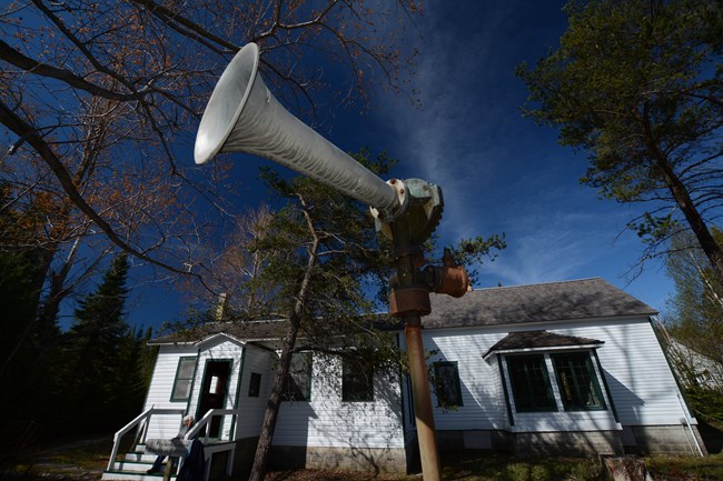 The original diaphone horn, seen outside the SMI Visitor Center A view looking up towards the sky at a grey steel funnel connected to a perpendicular pipe. A white building can be seen in the background.