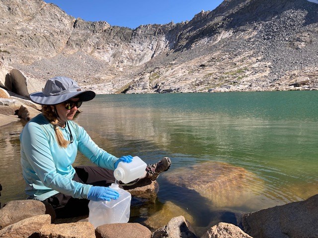 Woman sits at edge of mountain lake pouring sampled water from a plastic bottle into a large jug.