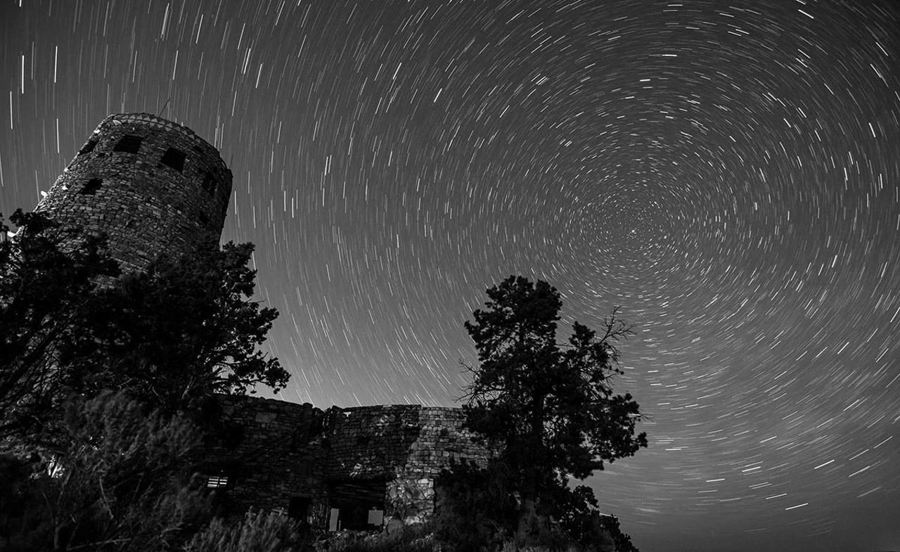 Stars over Desert View Watchtower A black and white image of the stars rotating around Polaris, a tall cylindrical building is in the foreground surrounded by trees.