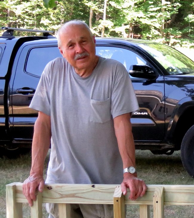 A man in a gray shirt stands with his hands resting on a wooden bike rack. There is a black truck behind him.