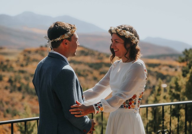Woman wearing flower-embroidered wedding dress and band of flowers in her hair holds her husband's arms and smiles at him, with view of mountains in background.