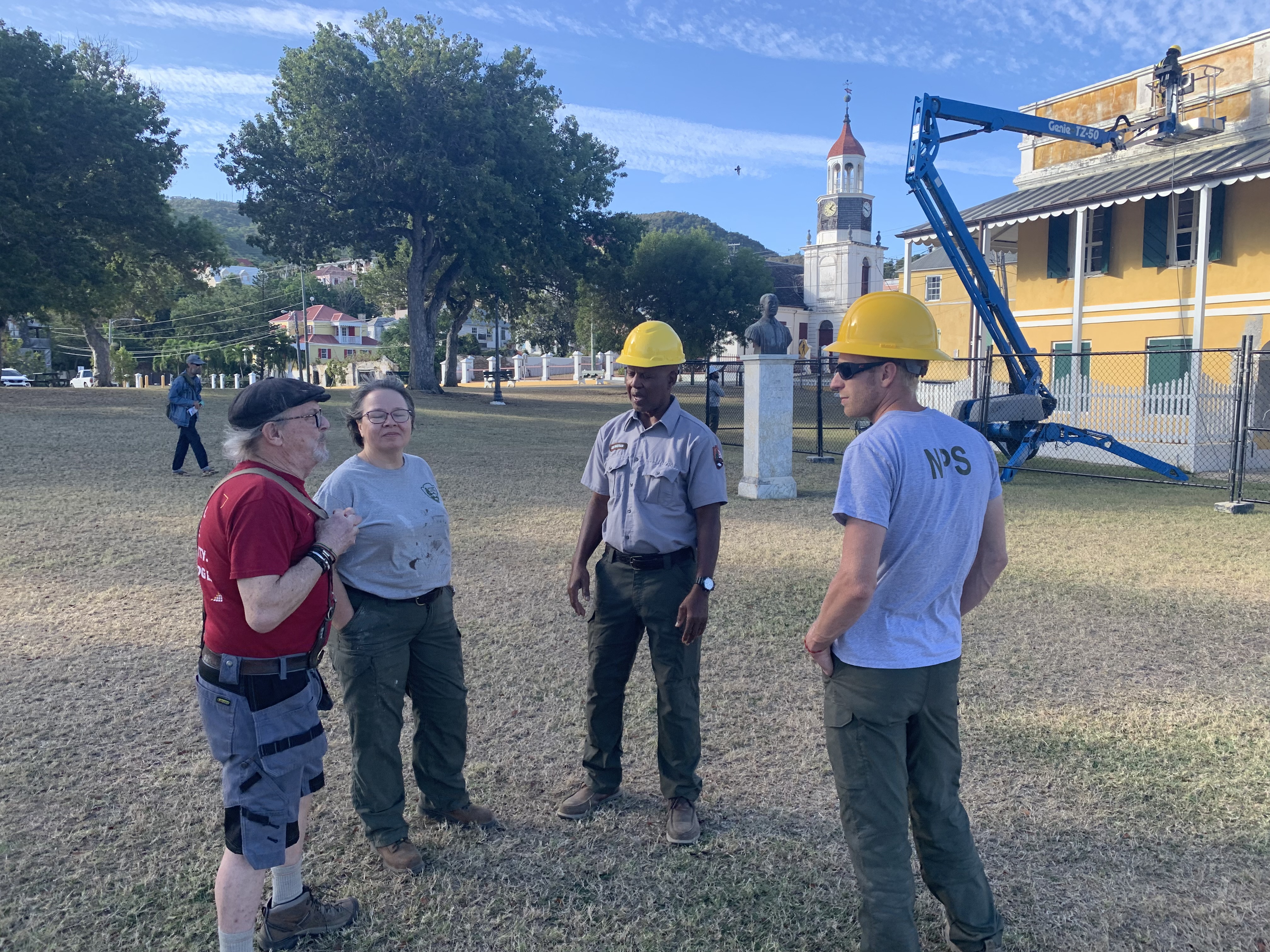 National Park Service employees stand in a semi-circle in the grassy foreground having a conversation as other staff members work on the Danish Customs House in the background.