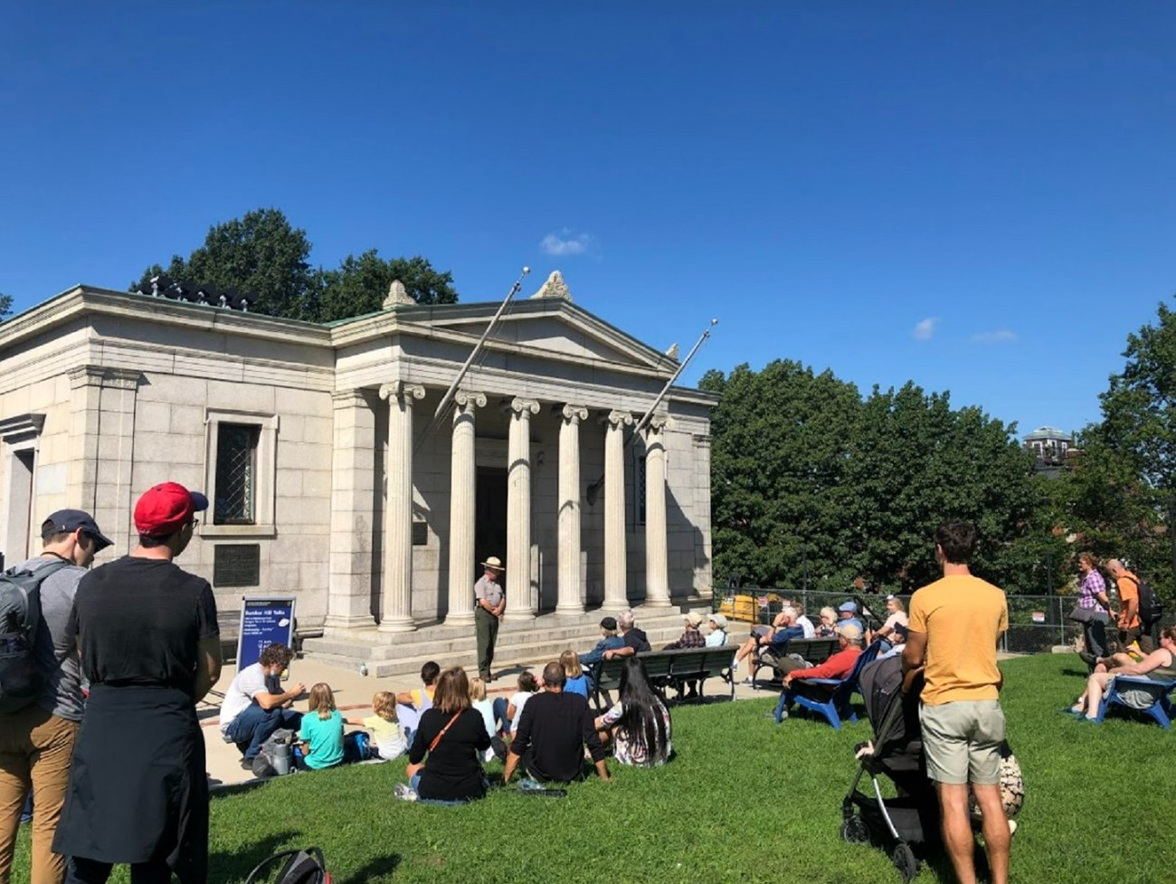 Ranger standing in front of a columned granite building and talking with a group of visitors who are dispersed around him.