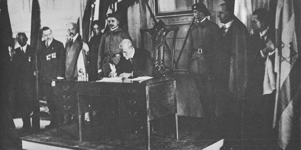 Black and white photo of a seated man signing a document while onlookers to his left and right watch.