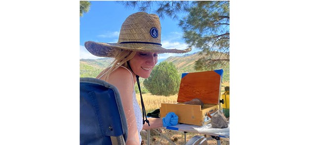 Woman in straw hat sits with painting tools in front of landscape with trees, grass, and mountains