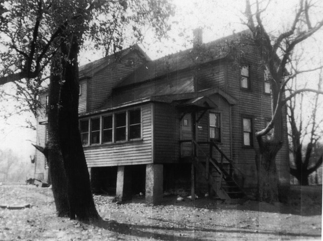 Black and white photo of a large, 2-story, wooden house with a complex roof, surrounded by tall trees. The front door leads into an enclosed porch with a row of 5 windows. The porch is raised up high on cement blocks.