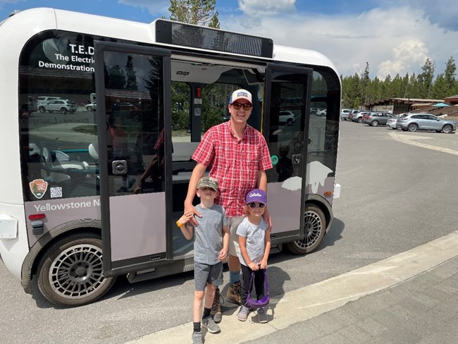 An adult and two children stand in front of an automated electric shuttle at Yellowstone National Park.