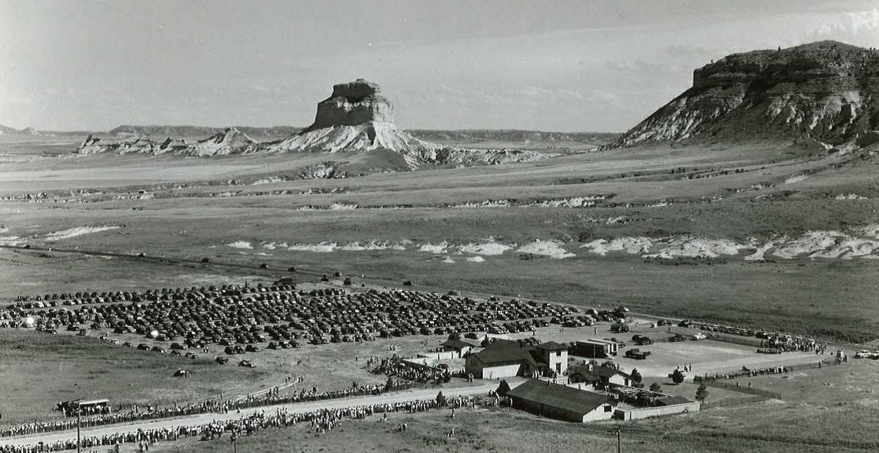 A black and white aerial photo of hundreds of cars parked in the prairie with sandstone bluff in the background.