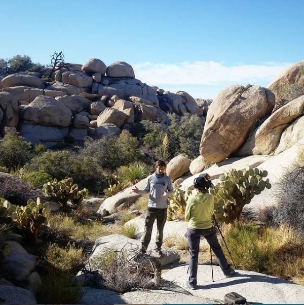 Videographer films a scientist explaining desert springs monitoring amidst desert vegetation and rocks.