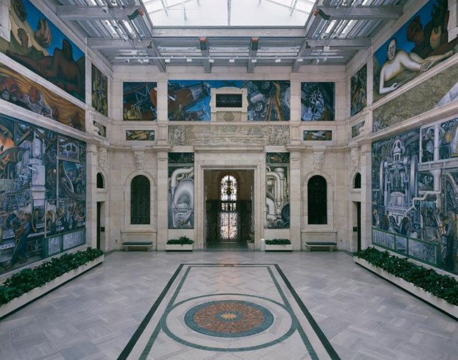 Detroit Industry mural The interior walls of a white marble building with a skylight and arched windows, are painted with murals of blues, greens, and browns, representing labor history.