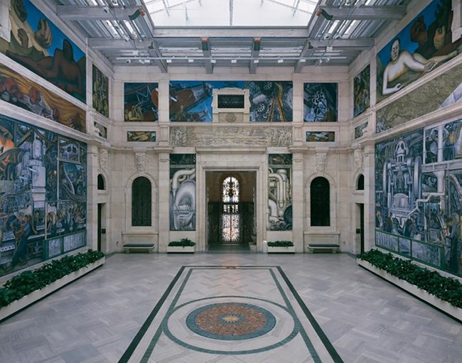 The interior walls of a white marble building with a skylight and arched windows, are painted with murals of blues, greens, and browns, representing labor history.