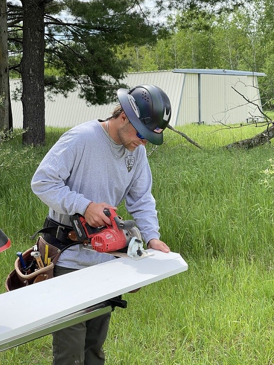 Devin Burkhart Worker wears a black hardhat with a brim, dark sunglasses and a grey national park service longsleeve shirt while looking down and cutting a plank of wood with a hand saw.