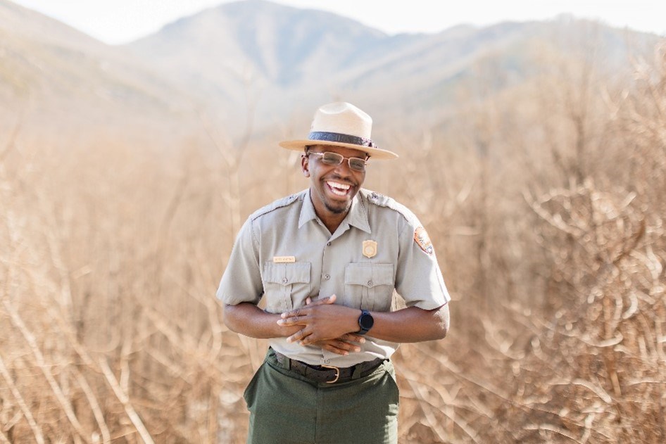 National Park Ranger Dexter Armstrong A smiling park ranger holding his hands near his chest and standing in a field. Rolling mountains are blurred in the distance.