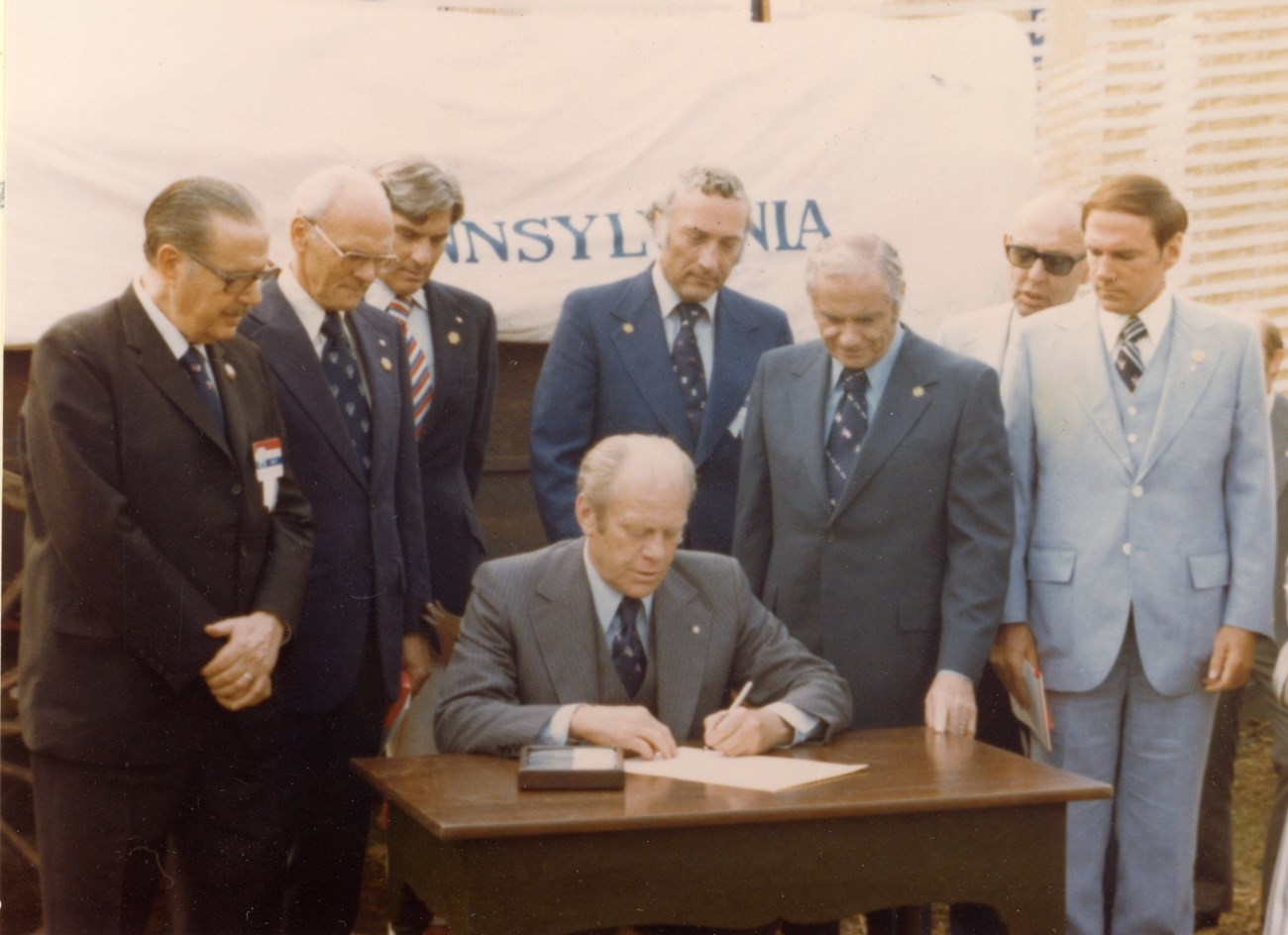 A group of men stand around president ford as he signs a document.