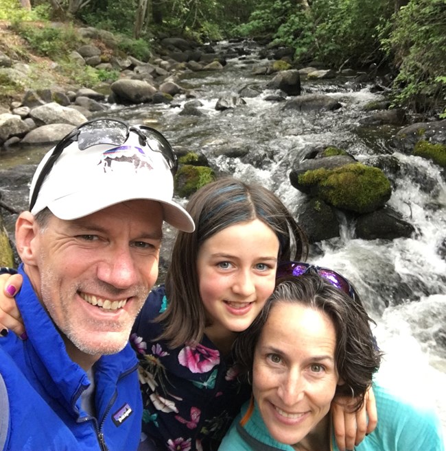 Dinger and family Man, woman and child hug each other in front of a stream.