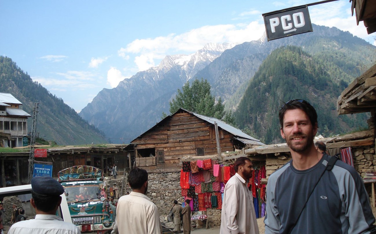 Dinger in Kashmir, Pakistan Smiling man on street of small village in Pakistan with colorful material hanging near a house and tall mountains in the background.