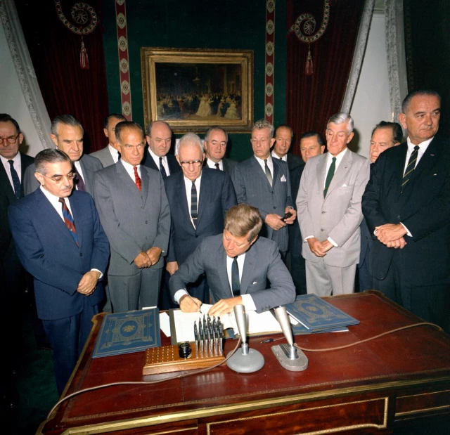 President John F. Kennedy signing the Nuclear Test Ban Treat. October 7, 1963. President JFK signs a paper on his desk, surrounded by old white men wearing suits—including vice president lyndon johnson.