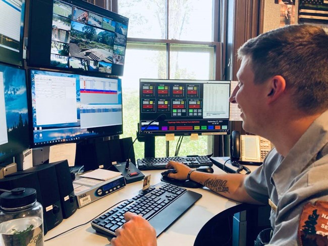 Dispatcher at Comm Ctr Uniformed ranger with short hair sits at a desk with at least five computer monitors; on the screens are colorful program interfaces and multiple video feeds.
