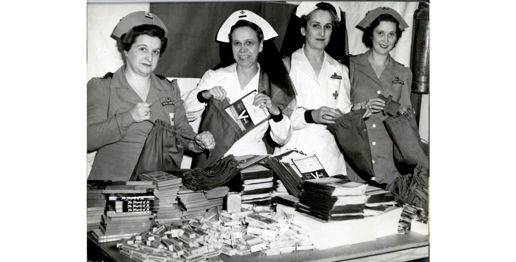Photo 2: Wichita Women making Ditty Bags Black and white photo of 4 white women wearing white or tan Red Cross uniforms and nurses' caps. They are putting booklets and other items into bags from a table in front of them.
