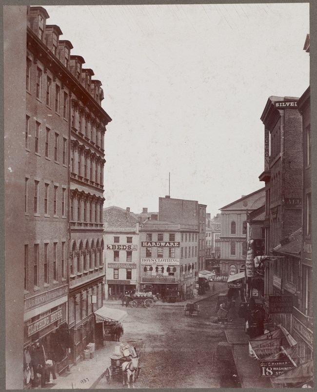 View down a street facing a small square. At the end are shops and people in horse drawn carriages