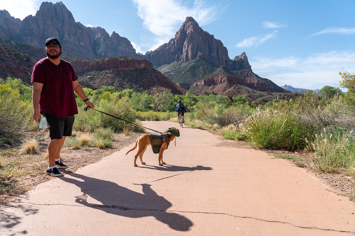 A man in a red shirt and shorts holds the leash of a dog on a paved path, a bike rider and views of Zion are in the background.